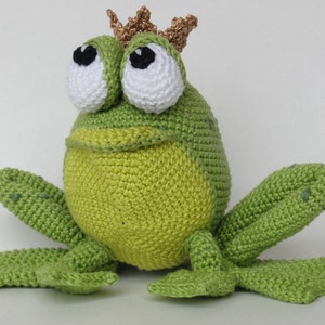 Peut inclure: Une grenouille en crochet verte portant une couronne dorée. La grenouille a de grands yeux blancs avec des pupilles noires et un large sourire.