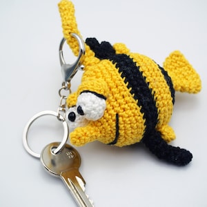 Puede incluir: Un llavero de abeja de crochet amarillo y negro con grandes ojos blancos. El llavero está unido a un llavero plateado y una llave plateada.
