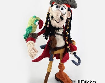 May include: A crocheted pirate doll with a dark gray hat, black hair, and a mustache. The doll wears a red coat, tan pants, and brown boots, with a hook hand and a small green parrot perched on the other arm. Text reads: © Ildikko www.ildikko.com