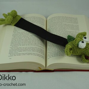 May include: A green crocheted dragon bookmark with large eyes and a pink tongue. The bookmark is attached to a black ribbon and is placed on a book.