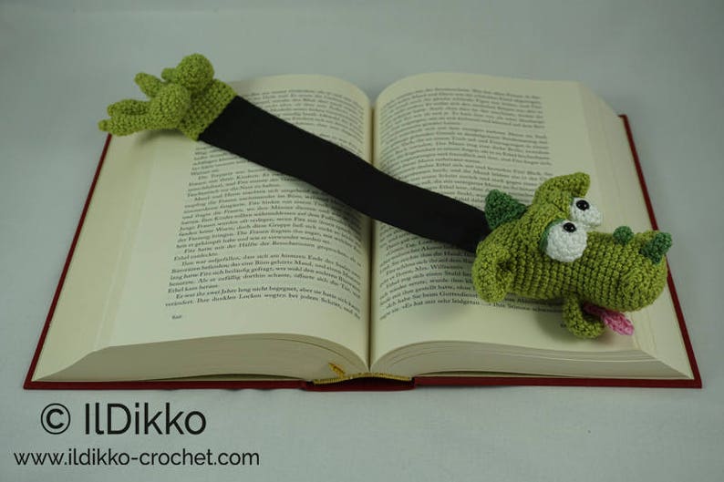 May include: A green crocheted dragon bookmark with large eyes and a pink tongue. The bookmark is attached to a black ribbon and is placed on a book.