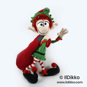 May include: A crocheted elf doll with red hair, a green tunic, and red and white striped leggings. The elf wears a green hat with a red pom-pom and carries a large red sack over one shoulder. The text "&copy; IlDikko www.ildikko.com" is visible.