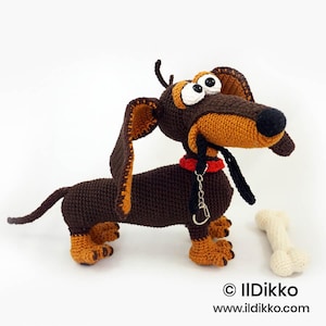 May include: A crocheted dachshund toy in dark brown and tan with large white eyes, a black nose, and a red collar featuring a silver metal keychain attachment. A small, off-white crocheted bone sits beside the dog. Text reads: © IlDikko www.ildikko.com