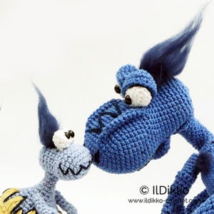 May include: Two blue crocheted dragon figures with large white eyes, black pupils, and dark blue tufts of hair on their heads. The larger dragon leans toward the smaller one. Text reads: &copy; ILDikko www.ildikko-crochet.com.