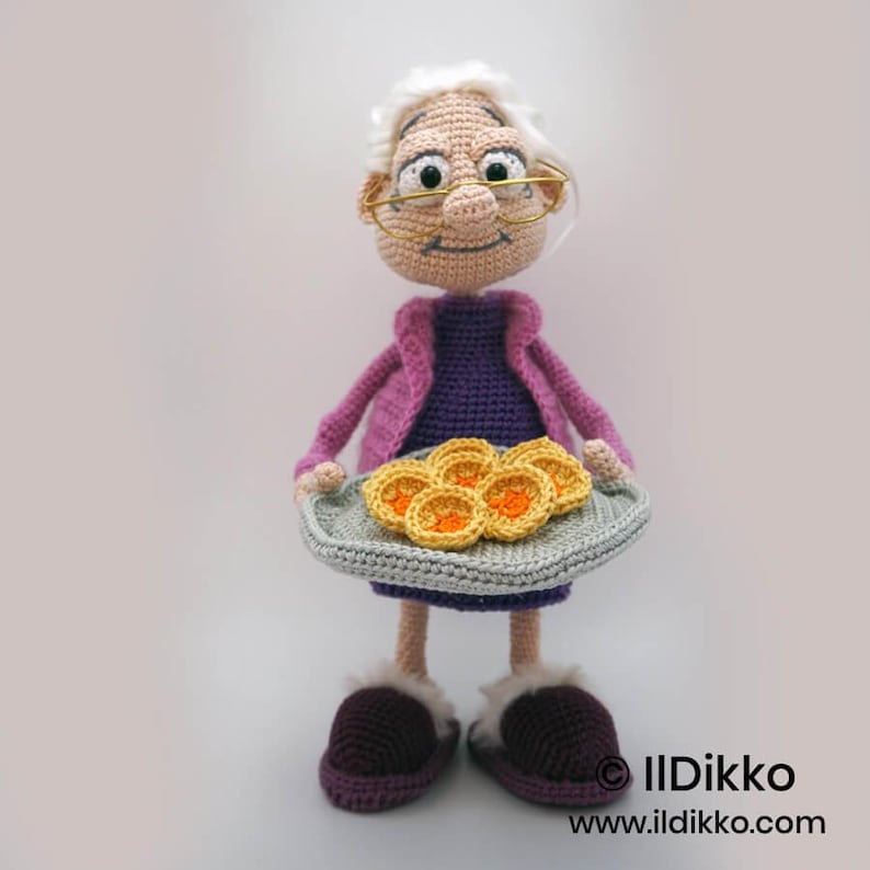 May include: A crocheted doll of a smiling elderly woman with glasses, white hair, and a purple dress, holding a tray of pancakes. She wears a pink jacket and purple slippers. The doll is handmade.