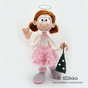 May include: A crocheted angel doll with auburn hair, a wire halo, and white wings. The doll wears a light gray top, a fluffy pink skirt, and gray shoes with pink trim. It holds a small, dark green crocheted Christmas tree with white beads.