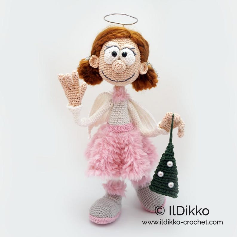 May include: A crocheted angel doll with auburn hair, a wire halo, and white wings. The doll wears a light gray top, a fluffy pink skirt, and gray shoes with pink trim. It holds a small, dark green crocheted Christmas tree with white beads.
