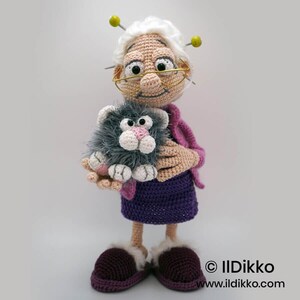 May include: A handmade crochet doll of a smiling elderly woman wearing glasses, holding a gray cat. The doll has white hair, a purple dress, and burgundy slippers. The cat has a pink nose and white paws. The doll is approximately 10 inches tall.