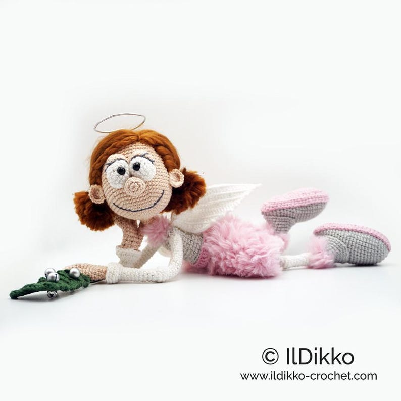 May include: A crocheted doll with auburn hair, a silver halo, and white wings. The doll wears a pink fuzzy skirt, grey and pink shoes, and holds a green crocheted leaf with silver bells. Text reads: &copy; ILDikko www.ildikko-crochet.com