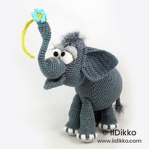 Puede incluir: Una figura de elefante tejida a ganchillo en color gris con grandes ojos blancos, un mechón de pelo gris en la cabeza y una larga trompa que sostiene un tallo amarillo con una flor azul claro. El elefante tiene detalles blancos en los pies.