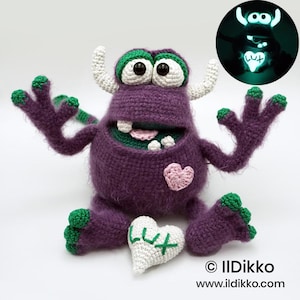 May include: A purple crochet monster doll with white horns, green accents on its hands and feet, and large black eyes. It features a pink heart patch on its chest and holds a white heart embroidered with the text LUX, which glows in the dark.