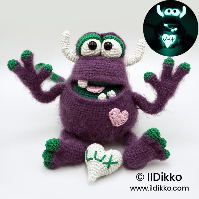 Puede incluir: Un mu&ntilde;eco monstruo de ganchillo morado con cuernos blancos, detalles verdes en manos y pies, y grandes ojos negros. Tiene un parche de coraz&oacute;n rosa en el pecho y sostiene un coraz&oacute;n blanco bordado con el texto LUX que brilla en la oscuridad.