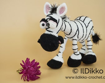 May include: A crocheted zebra toy with black and white stripes, large eyes, and a black nose. The zebra is standing on a light brown surface and looking down at a pink flower.