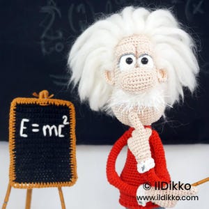 May include: A crocheted doll of a scientist with wild white hair, a mustache, and a red shirt, standing next to a small black chalkboard with the white text E=mc2. The figure has a beige complexion, large eyes, and a hand raised to its chin.