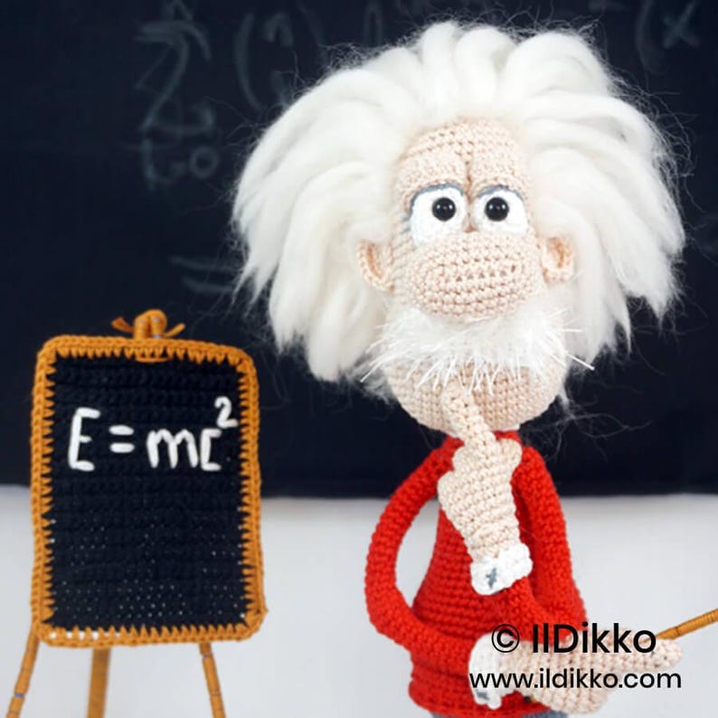 May include: A crocheted doll of a scientist with wild white hair, a mustache, and a red shirt, standing next to a small black chalkboard with the white text E=mc2. The figure has a beige complexion, large eyes, and a hand raised to its chin.