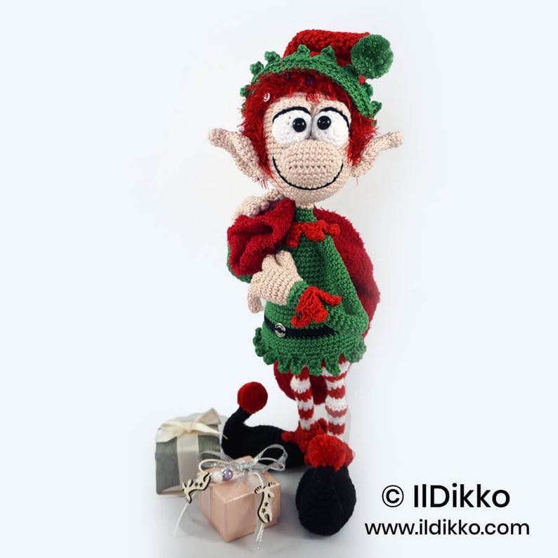 May include: A crocheted elf doll with red hair, a green and red outfit, and a matching hat, carrying a red sack. The elf has large eyes and pointed ears. Two small gift boxes with ribbons are placed at the base. Text reads: &copy; IIDikko www.ildikko.com