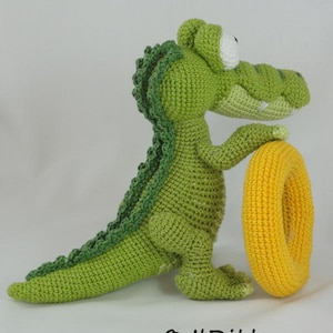 May include: A green crocheted crocodile toy with a textured back, large white eyes, and a light green snout, standing upright while holding a bright yellow crocheted ring. The text "&copy; ILDikko www.ildikko-crochet.com" is printed at the bottom.