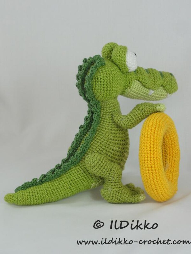May include: A green crocheted crocodile toy with a textured back, large white eyes, and a light green snout, standing upright while holding a bright yellow crocheted ring. The text "&copy; ILDikko www.ildikko-crochet.com" is printed at the bottom.