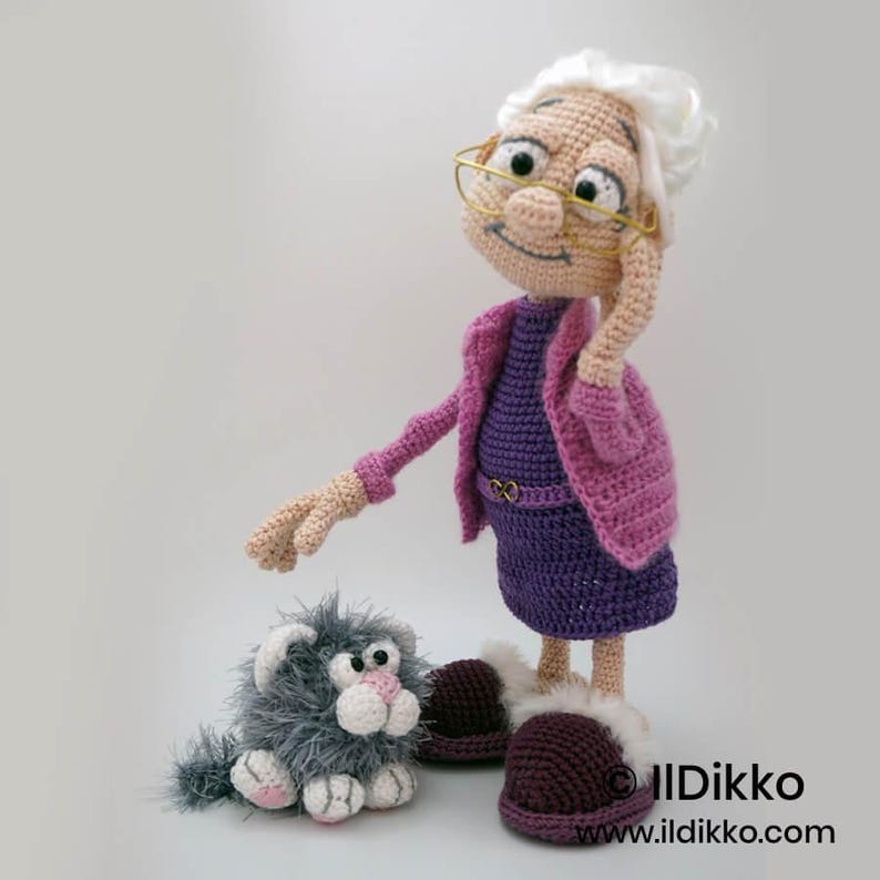 May include: A handmade crochet doll of an elderly woman wearing a purple dress and pink jacket, with glasses and white hair. A small gray cat is seated next to her. The doll is wearing burgundy slippers.