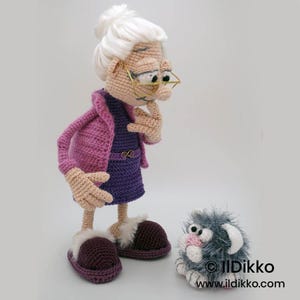 May include: A handmade crochet doll of an elderly woman wearing a purple dress, pink jacket, and slippers. The doll has white hair, glasses, and is deep in thought. A small, gray, fluffy crochet creature sits nearby. The doll is approximately 12 inches tall.