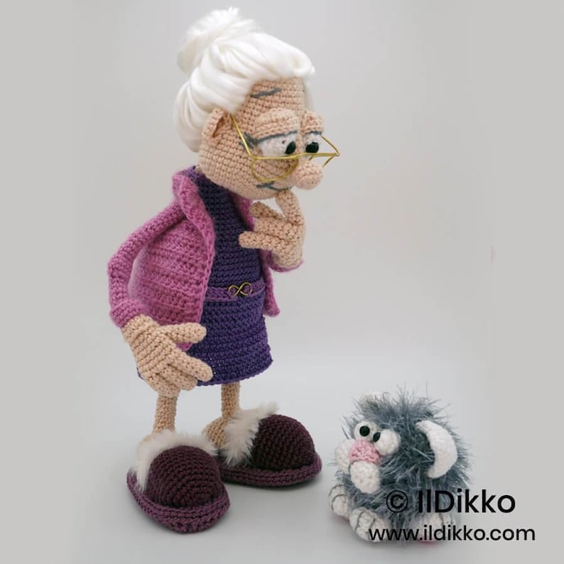 May include: A handmade crochet doll of an elderly woman wearing a purple dress, pink jacket, and slippers. The doll has white hair, glasses, and is deep in thought. A small, gray, fluffy crochet creature sits nearby. The doll is approximately 12 inches tall.