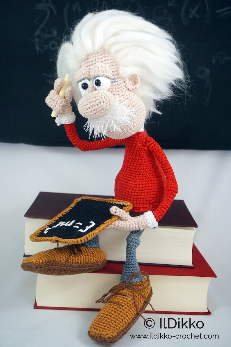 May include: A crocheted doll of an elderly scientist with wild white hair, wearing a red long-sleeved shirt and gray trousers. The figure holds a small piece of chalk and sits on two stacked books, holding a black chalkboard with E=mc2 written on it.