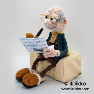 May include: A crocheted doll of an elderly man wearing glasses, reading a newspaper titled "DER SPIEGETTI". The doll is dressed in a green jacket, yellow shirt, and brown pants. He is seated on a beige cushion, with a pipe and tobacco tin nearby.