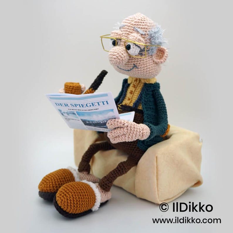 May include: A crocheted doll of an elderly man wearing glasses, reading a newspaper titled "DER SPIEGETTI". The doll is dressed in a green jacket, yellow shirt, and brown pants. He is seated on a beige cushion, with a pipe and tobacco tin nearby.