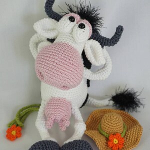 May include: A crocheted cow toy with black and white fur, pink nose, and large eyes. The cow is wearing a brown crocheted hat with a flower. The toy is sitting on a white surface. &copy; Ildikko www.ildikko-crochet.com