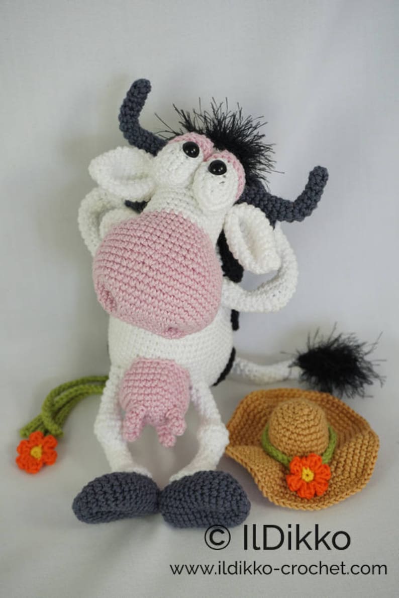 May include: A crocheted cow toy with black and white fur, pink nose, and large eyes. The cow is wearing a brown crocheted hat with a flower. The toy is sitting on a white surface. &copy; Ildikko www.ildikko-crochet.com