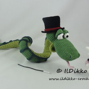 Puede incluir: Una serpiente de crochet verde que lleva un sombrero de copa negro con una banda roja, mirando a un conejo de crochet blanco. La serpiente tiene un cuerpo largo y sinuoso y ojos grandes y negros. El conejo tiene orejas rosas y ojos negros. La imagen incluye el texto "@ ILdikko" y "www.ildikko-crochet.com".