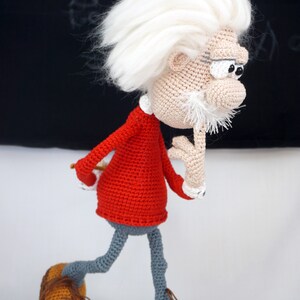 May include: A crocheted doll of an elderly person with wild white hair, a white mustache, and thick eyebrows. The doll wears a red long-sleeved shirt, gray trousers, and brown shoes with laces. One hand is raised to the chin in a thoughtful pose.