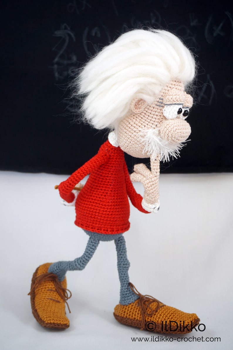 May include: A crocheted doll of an elderly person with wild white hair, a white mustache, and thick eyebrows. The doll wears a red long-sleeved shirt, gray trousers, and brown shoes with laces. One hand is raised to the chin in a thoughtful pose.