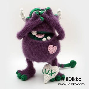 Puede incluir: Un monstruo de ganchillo morado con detalles verdes, cuernos blancos y un parche de coraz&oacute;n rosa. El monstruo se cubre los ojos con las manos y sostiene un coj&iacute;n blanco en forma de coraz&oacute;n con el texto bordado en verde LUX. Tiene cola verde.