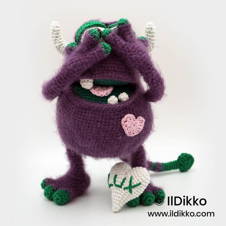 Puede incluir: Un monstruo de ganchillo morado con detalles verdes, cuernos blancos y un parche de coraz&oacute;n rosa. El monstruo se cubre los ojos con las manos y sostiene un coj&iacute;n blanco en forma de coraz&oacute;n con el texto bordado en verde LUX. Tiene cola verde.