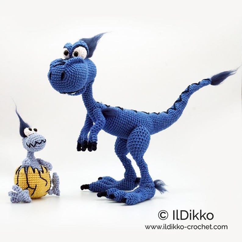 May include: A large blue crocheted dinosaur with black claws and a tuft of hair, standing next to a smaller light blue crocheted dinosaur emerging from a yellow cracked egg. The text ILDikko and www.ildikko-crochet.com appears at the bottom.