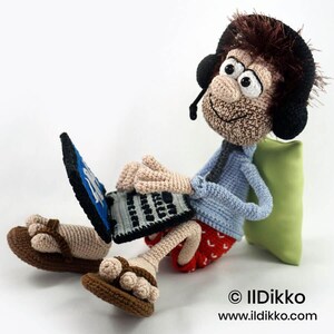 May include: A crocheted doll with brown hair wearing black headphones, a light blue shirt, and red patterned shorts. The doll sits while using a black and blue laptop, wearing brown sandals, and leaning against a small light green pillow.