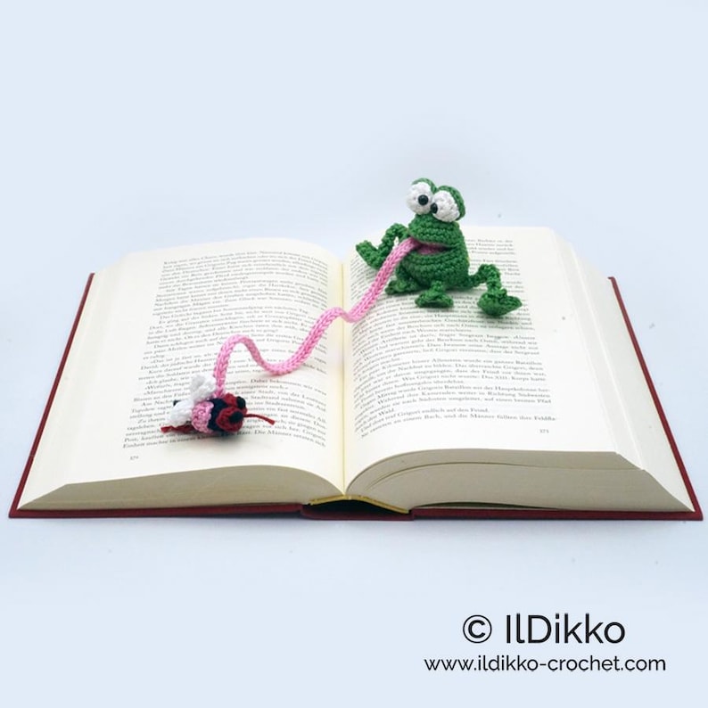 May include: A green crocheted frog bookmark with a pink crocheted tongue catching a crocheted fly. The bookmark is placed in an open book.