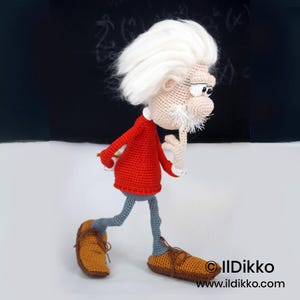 May include: A crocheted doll of an elderly person with wild white hair, a white mustache, and a red long-sleeved shirt. The figure has grey trousers, brown shoes, and a beige complexion, with one hand touching the chin in a thoughtful pose.
