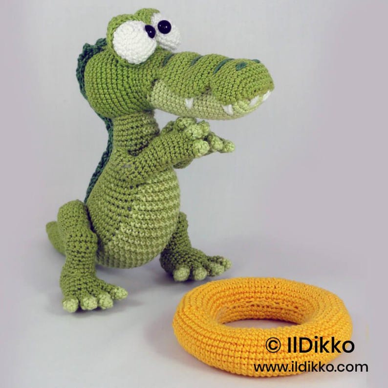 May include: A green crocheted alligator toy with large white eyes and black pupils, standing upright with small claws. Beside it lies a round, yellow crocheted ring. The text "&copy; Ildikko www.ildikko.com" is printed in the lower right corner.