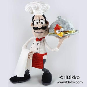 May include: A crocheted chef doll wearing a white toque, a white jacket with black cross-stitch details, a red apron, and black shoes. The doll holds a white cloth in one hand and a grey crocheted serving cloche covering a chicken dish in the other.