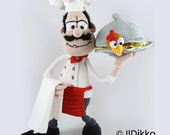 May include: A crocheted chef doll wearing a white toque, a white jacket with black cross-stitch details, a red apron, and black shoes. The doll holds a white cloth in one hand and a grey crocheted serving cloche covering a chicken dish in the other.