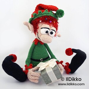 May include: A crocheted elf doll with red hair, a green and red hat with a green pom-pom, and a green outfit. The elf has large eyes, pointed ears, and black curled shoes. It holds a grey gift box with a white ribbon. Text reads: &copy; IlDikko www.ildikko.com
