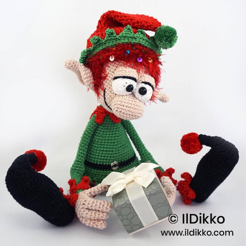 May include: A crocheted elf doll with red hair, a green and red hat with a green pom-pom, and a green outfit. The elf has large eyes, pointed ears, and black curled shoes. It holds a grey gift box with a white ribbon. Text reads: &copy; IlDikko www.ildikko.com