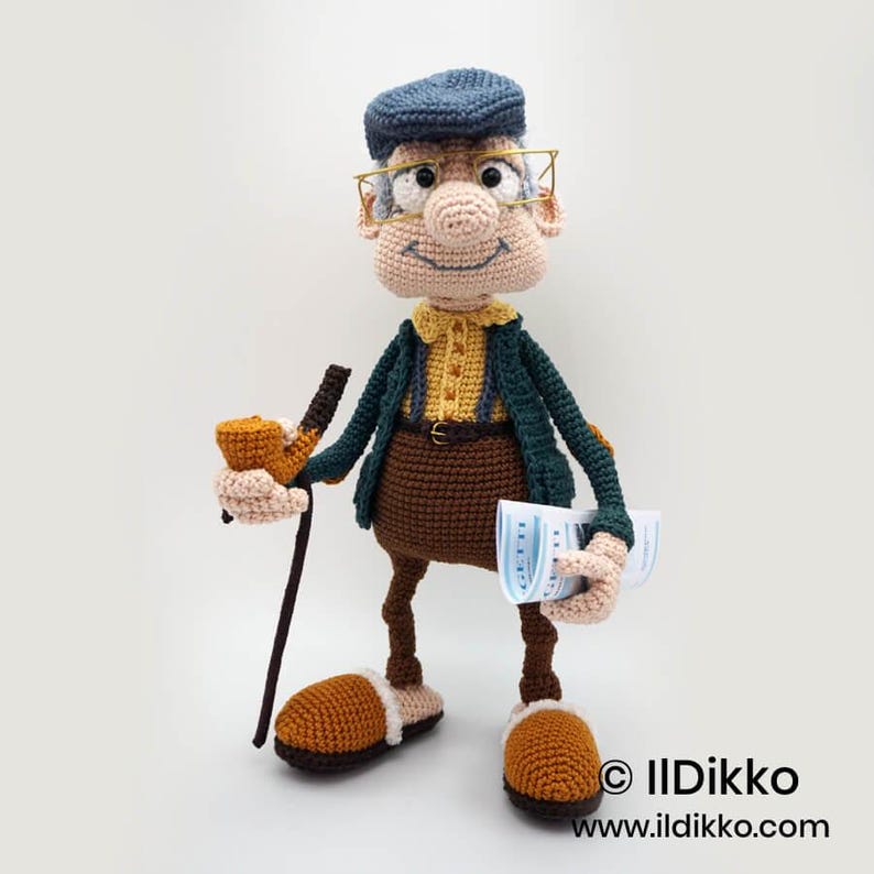 May include: A crocheted figure of an elderly man wearing a blue cap, glasses, and a green jacket over a yellow shirt. He holds a pipe and a walking stick in one hand and a newspaper in the other. The figure wears brown pants and slippers.