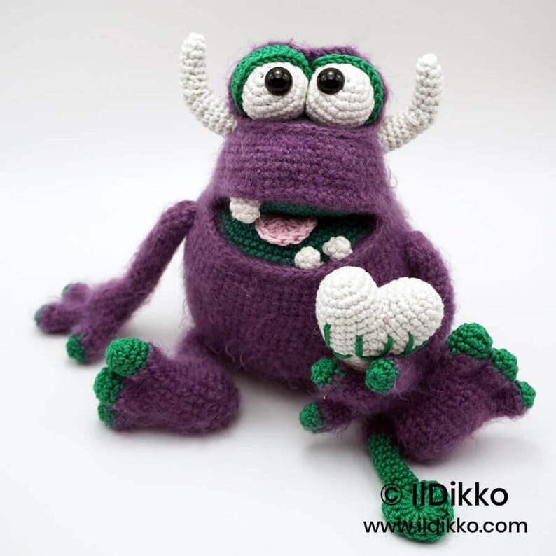 Puede incluir: Un juguete de monstruo tejido a ganchillo de color morado con dos cuernos blancos, ojos grandes blancos con pupilas negras y detalles verdes en pies y manos. El monstruo tiene la boca abierta mostrando dientes blancos y una lengua rosa, sosteniendo un coraz&oacute;n.