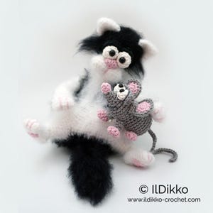 Puede incluir: Un gato de crochet blanco y negro sosteniendo un ratón de crochet gris con detalles rosas. El gato tiene grandes ojos negros y una cola esponjosa.