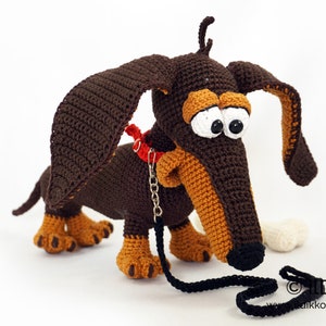May include: A crocheted dachshund dog with large, expressive eyes, brown and tan coloring, and a red collar. The dog has a black leash and a white bone. The dog is handmade.
