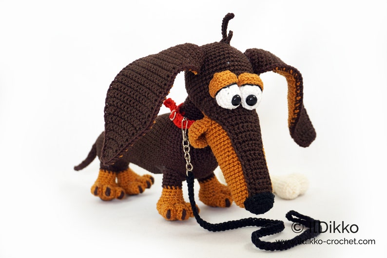May include: A crocheted dachshund dog with large, expressive eyes, brown and tan coloring, and a red collar. The dog has a black leash and a white bone. The dog is handmade.