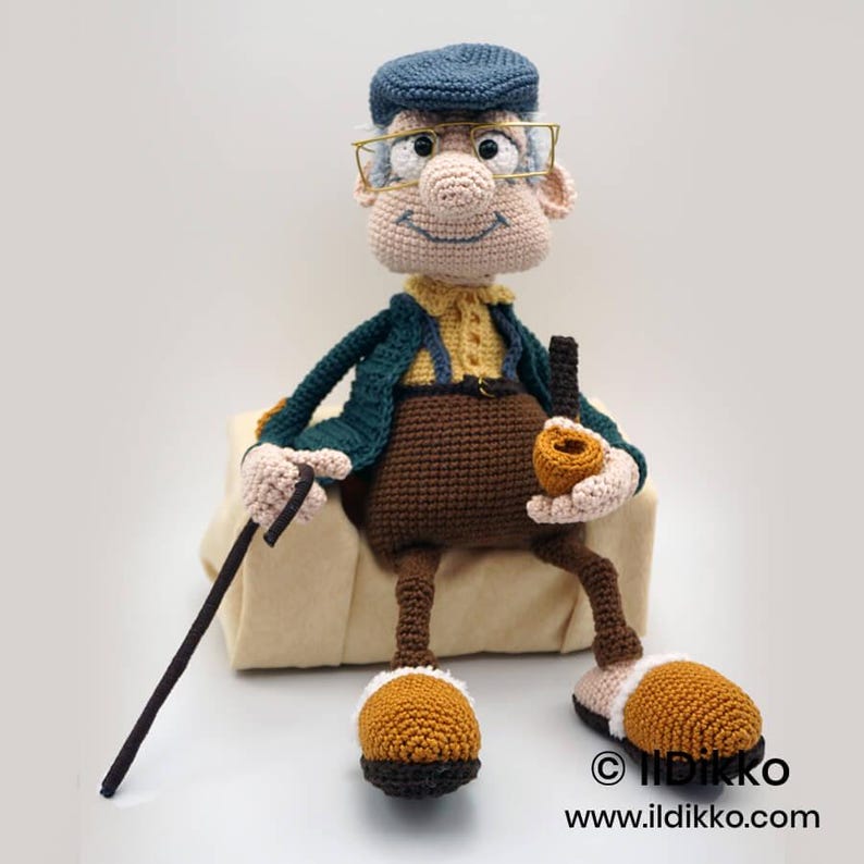 May include: A handmade crochet doll of an elderly man, wearing a blue cap, glasses, and a green jacket over a yellow shirt. He holds a pipe and a cane. The doll has brown pants and shoes with white trim. The doll is sitting on a beige cushion.